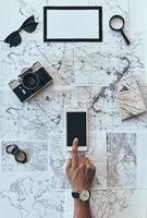 I want to visit every place. Close up top view of man using smart phone with sunglasses, photo camera, compass, magnifying glass and passport lying on map around