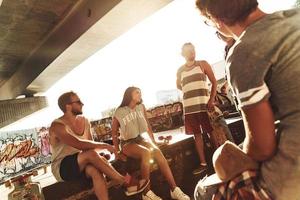 Enjoying sunny day. Group of skater friends hanging out together while resting under the bridge outdoors photo