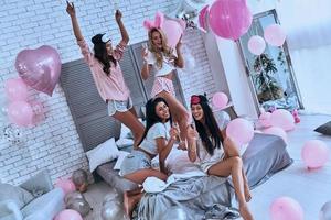 la fiesta es imposible sin la vista superior de champán de cuatro mujeres sonrientes jóvenes juguetonas en pijama uniéndose mientras tienen una fiesta de pijamas en el dormitorio con globos por todo el lugar foto