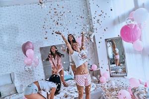 tan felices cuatro atractivas mujeres jóvenes en pijama sonriendo y gesticulando mientras saltan en el dormitorio con confeti volando por todas partes foto