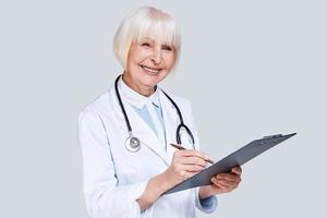 New prescription. Beautiful senior woman in lab coat looking at camera and writing something down while standing against grey background photo