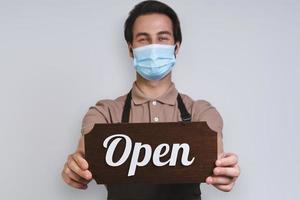 Handsome young man in apron wearing protective face mask and holding open sign while standing against gray background photo