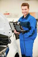 Ahora sé el problema. joven confiado trabajando en una laptop especial y sonriendo mientras está de pie en un taller cerca de un auto foto