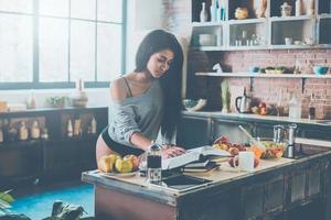 Reading cookbook. Beautiful young mixed race woman reading cookbook while standing in kitchen at home photo