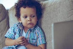 That was unexpected Little African baby boy looking at his laptop and looking confused while sitting on the couch at home photo