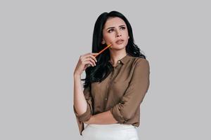 Thoughtful young woman in smart casual wear touching her cheek with pencil and looking away while standing against grey background photo