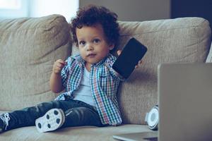 Can you dial with dad Little African baby boy giving his smartphone to you and looking at camera while sitting on the couch at home photo