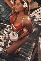 naturalmente bello. vista superior de una atractiva joven en bikini rojo sosteniendo un cóctel de coco mientras camina por los escalones de madera al aire libre foto
