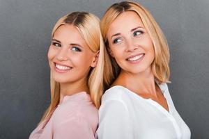 Tender moments with mother. Beautiful mother and her daughter smiling while standing back to back against grey background photo