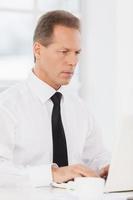 empresario en el trabajo. hombre maduro confiado en camisa y corbata usando computadora mientras está sentado en su lugar de trabajo foto