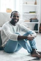 Enjoying weekend at home Handsome young African man in smart casual clothes looking at camera and smiling while sitting on the carpet at home photo