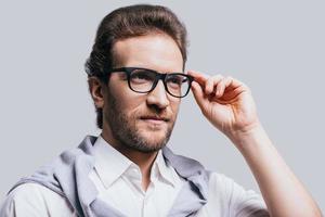 Looking in the future Handsome young man in smart casual clothes adjusting his eyeglasses and looking away while standing against grey background photo