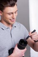 Photographer cleaning the lens. Handsome young man in polo shirt holding digital camera and cleaning the lens with lens brush while standing in studio photo