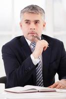 Waiting for inspiration. Thoughtful senior man in formalwear holding pen on chin and looking away while sitting at his working place photo