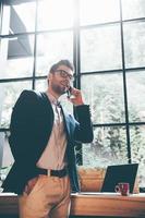 Good news from colleague. Low angle view of confident young man talking on the mobile phone and smiling while standing in front of the big window in office or cafe photo