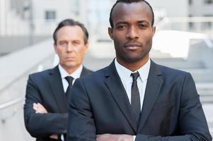 Confident business experts. Confident African man in formalwear keeping arms crossed and looking at camera while mature businessman standing behind him photo