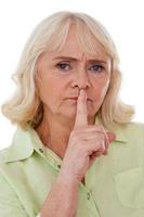 Keep silence Serious senior woman holding finger on mouth and looking at camera while standing isolated on white background photo