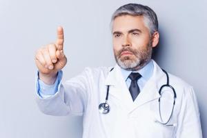 Doctor working on transparent wipe board. Mature grey hair doctor touching transparent wipe board with finger while standing against grey background photo