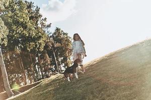 disfrutando del día de verano. toda la longitud de una linda niña jugando con su perro mientras corre al aire libre foto