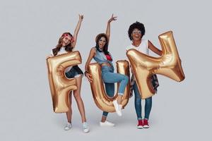 longitud total de tres atractivas mujeres jóvenes gesticulando y sonriendo mientras están de pie contra el fondo gris foto