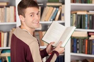 Enjoying his favorite book. Cheerful young man holding a book and looking over shoulder while standing at the library photo