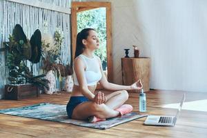 Beautiful young woman in sports clothing practicing yoga on patio with laptop laying near her photo