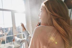 Rear view of beautiful young woman talking on her smart phone and smiling while sitting near the window at home photo