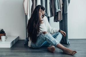 Hard choice.  Thoughtful young woman in casual wear sitting on the floor at home and holding hand in hair with clothes hanging in the background photo