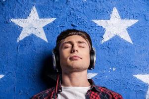 Enjoying music. Low angle view of young man in headphones keeping eyes closed while standing against background photo