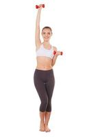 Beautiful and sporty. Full length of beautiful young woman in sports clothing training with dumbbells and smiling while standing isolated on white photo