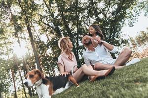 Momentos de alegría en familia. feliz familia joven de tres con perro sonriendo mientras se sienta en el césped en el parque foto