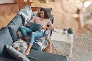 Top view of relaxed young woman in casual clothing using laptop and smiling while spending time at home photo