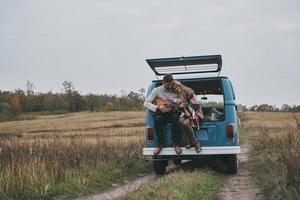 ella es su musa. un joven apuesto tocando la guitarra para su bella novia mientras se sienta en el maletero de una mini furgoneta azul de estilo retro