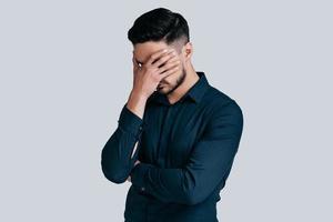 Feeling depressed.  Exhausted young man covering face with hand while standing against grey background photo