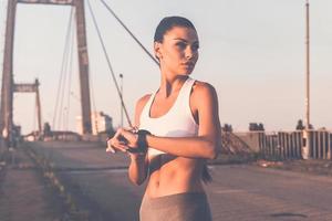 Ready to run. Beautiful young woman in sports clothing wearing smartwatch and looking away while standing on the bridge photo