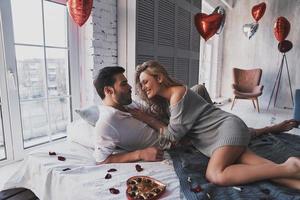 Sweet candies in bed.  Beautiful young couple eating chocolate candies and smiling while lying on the bed at home photo