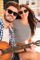 Spending carefree time together. Smiling young loving couple bonding to each other and looking at camera while sitting outdoors with guitar photo