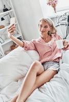 Sharing photos. Top view of attractive young woman taking selfie and smiling while sitting on bed at home photo