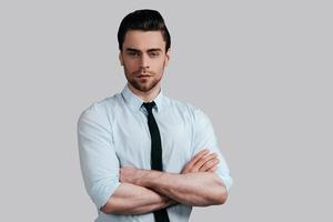 Confident and handsome. Handsome young man in white shirt and tie keeping arms crossed and looking at camera while standing against grey background photo