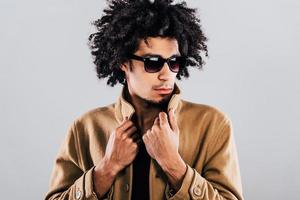 Not afraid of could weather. Close-up of confident young African man in sunglasses looking away and adjusting his coat while standing against grey background photo