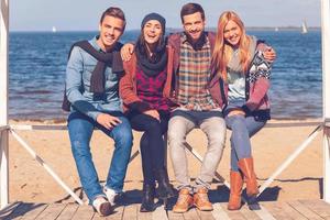 mejores amigos por siempre. longitud completa de cuatro jóvenes felices que se unen entre sí y sonríen mientras están sentados en la playa foto