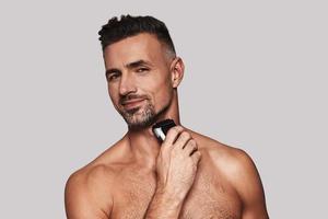 Everyday routine. Charming young man shaving and smiling while standing against grey background photo