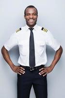 piloto confiado. piloto africano confiado en uniforme sosteniendo la mano en la cadera y sonriendo mientras está de pie contra el fondo gris foto