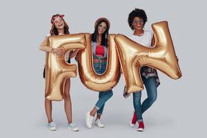 Full length of three attractive young women carrying balloons and smiling while standing against grey background photo