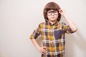 hipster joven serio. niño pequeño con anteojos cogido de la mano en la cadera y ajustando sus gafas mientras está de pie contra un fondo gris foto