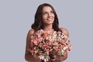 chica encantadora. mujer joven atractiva con ramo de flores mirando hacia otro lado y sonriendo mientras está de pie contra el fondo gris foto