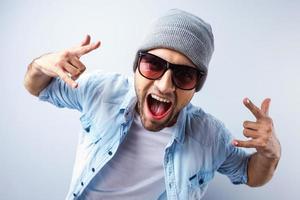 fresco y moderno. vista superior de un apuesto joven con sombrero y gafas de sol gesticulando y haciendo muecas mientras se enfrenta a un fondo gris foto