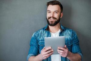 Smart technology makes everything easier. Happy young man holding digital tablet and smiling at camera while standing against grey background photo