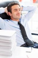 Businessman day dreaming. Handsome young beard man in shirt and tie holding hands behind head and smiling while sitting at the table with stack of paperwork laying on it photo