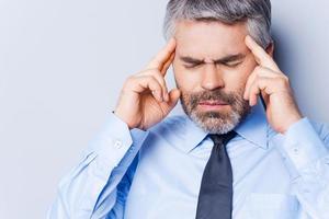 Suffering from headache. Depressed mature man in shirt and tie touching head with fingers and keeping eyes closed while standing against grey background photo